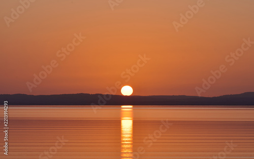 Beautiful golden sunset over calm coastal waters, with the setting sun casting a bright reflection across the sea and a dark distant shoreline beneath a warm orange sky.