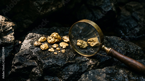 A moody, low-key of raw gold nuggets resting on dark, textured volcanic rock, viewed through a vintage brass magnifying glass with a wooden handle