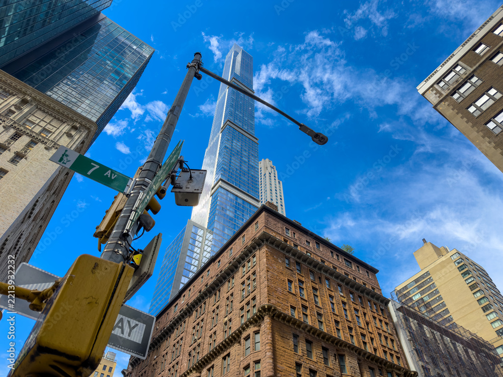 Naklejka premium A streetlight crosses the view of a skyscraper above a Manhattan corner. A yellow signal sits below green signage as clouds pass between glass and brick facades.