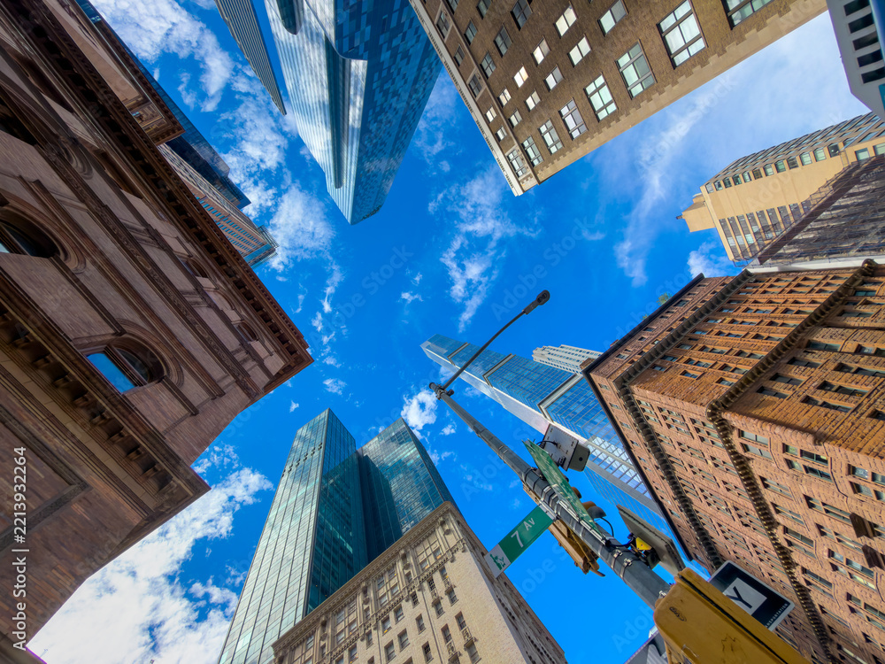 Fototapeta premium A ring of Manhattan buildings encloses the sky above a streetlight. The upward view places glass towers and masonry corners around clouds and green signage.