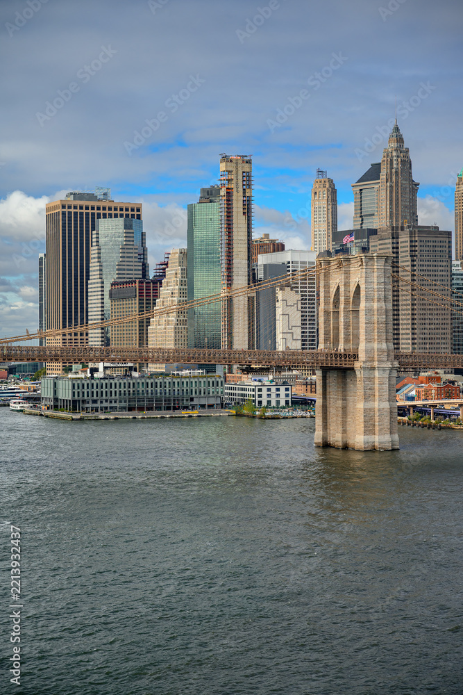 Fototapeta premium A stone tower of Brooklyn Bridge stands beside the Manhattan skyline. Suspension cables cross glass facades above a pier building and gray river water under clouds.
