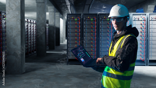 Energy storage facility, electrical engineer uses a laptop to monitor industrial battery racks, power distribution and high-voltage storage equipment.