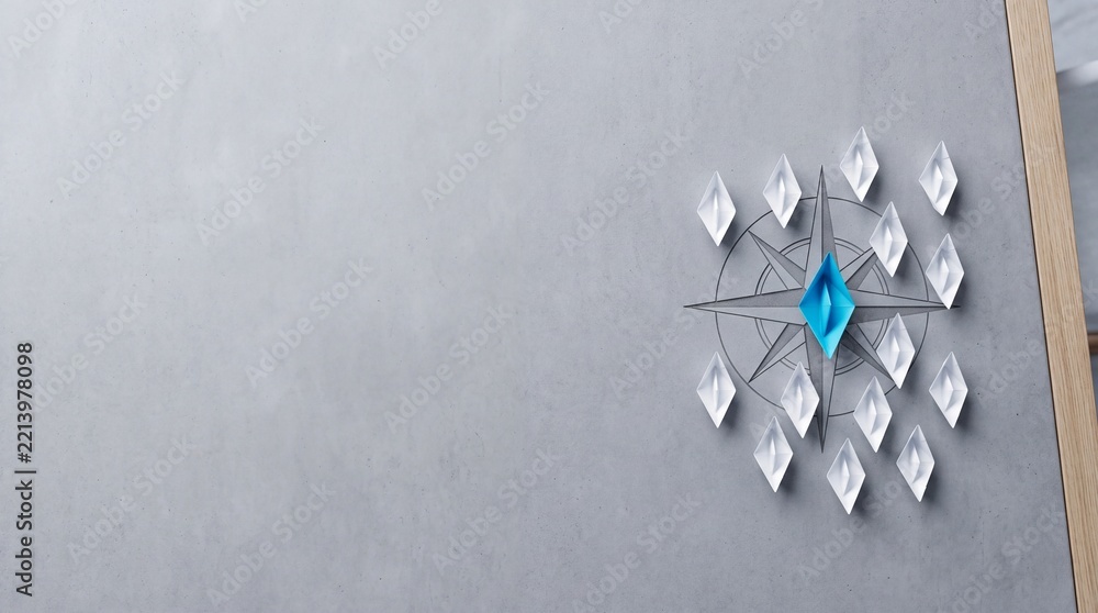 Fototapeta premium Conceptual image of a unique blue origami paper boat leading white boats on a compass rose symbol, representing leadership, vision, and business direction with ample negative space on the left