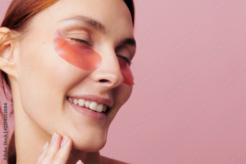 Obraz premium An ecstatic woman smiles with closed eyes, touching her chin in a tight crop. Hydrogel patches under her eyes create a vibrant self-pampering concept for beauty content.