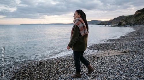 Thoughtful young woman walking alone along a pebbled beach by the sea on an overcast day