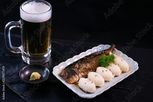 A ready-to-eat meal set consisting of rice balls and grilled mackerel, with a mug of beer arranged side-by-side on a black stone surface; a Japanese-style meal.