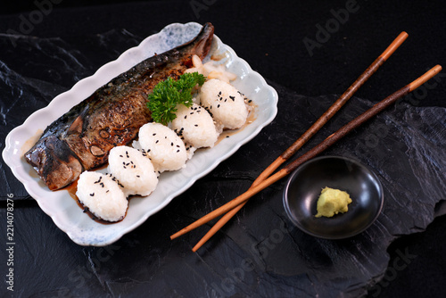 A ready-to-eat meal set featuring a rice ball and grilled mackerel, accompanied by chopsticks and wasabi in a small cup placed nearby. Japanese cuisine. View from above
