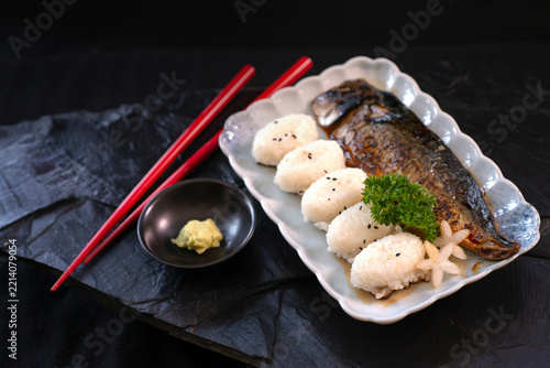 A ready-to-eat meal set consisting of a rice ball and grilled mackerel, accompanied by chopsticks and wasabi in a small cup, arranged side-by-side on a black stone surface; a Japanese-style meal.