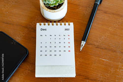 December 2027 desk calendar with pen, plant, and mobile phone on wooden background. Time management, planning, and schedule concept.