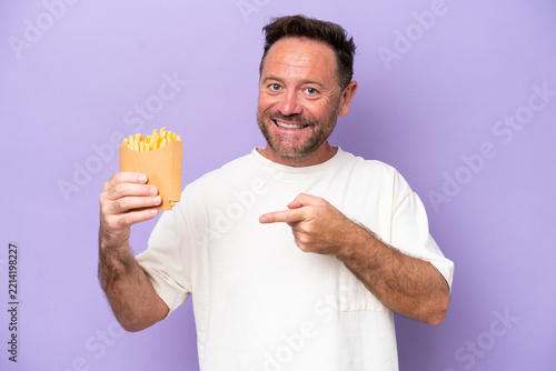 Middle age caucasian man holding fried chips isolated on purple bakcground pointing finger to the side