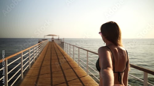 Woman stands on a wooden pier, gazing at the sea. Sunlight bathes her back as she looks out at the horizon. The calm waters stretch ahead, reflecting the soft glow. She holds her own hand