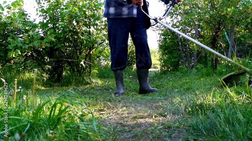 Person mows grass with handheld trimmer. Greenery surrounds the path. Grass blades fly upward with each cut. Sunlight glints off the trimmer. Routine garden maintenance captured