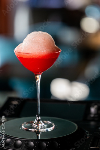 Red cocktail in a coupe glass topped with thick foam, resting on a DJ turntable in a dimly lit bar.