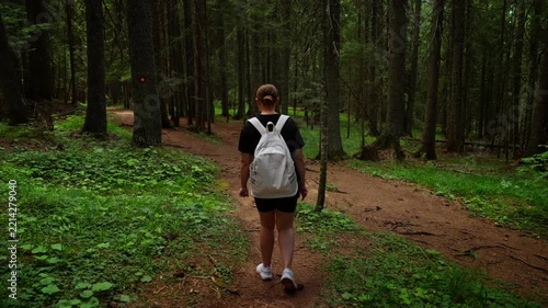 Woman with white backpack walking along peaceful forest trail, exploring tranquil woodland path surrounded by tall trees, green foliage, and soft natural light