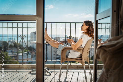Happy young woman sitting on balcony chair with barefoot legs on table and looking at smartphone screen.