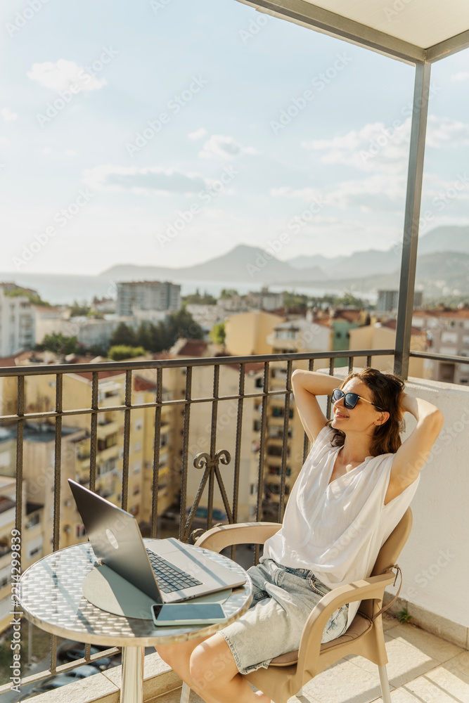 Fototapeta premium Relaxed young woman sitting on balcony with laptop and hands behind head enjoying city view.