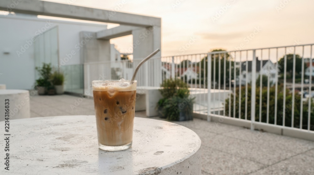 Fototapeta premium Refreshing iced latte in a glass with a metal straw on a modern rooftop terrace at sunset