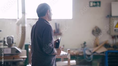 Male carpenter holding a digital tablet while walking inside his modern workshop. He is checking the quality of his wooden products