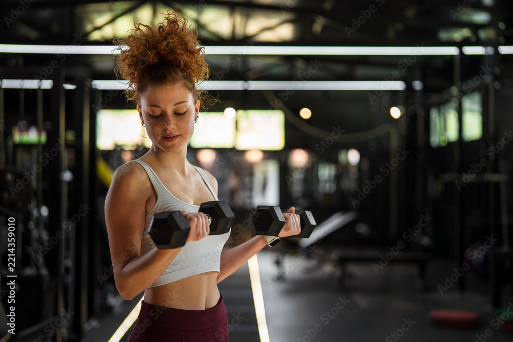 Fototapeta premium Young woman lifting dumbbells performing bicep curls in gym