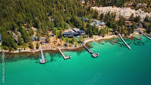 Cottages and docks line a forested shore at Lake Tahoe. Gray roofs sit among green conifers above turquoise water with narrow piers in an elevated view.