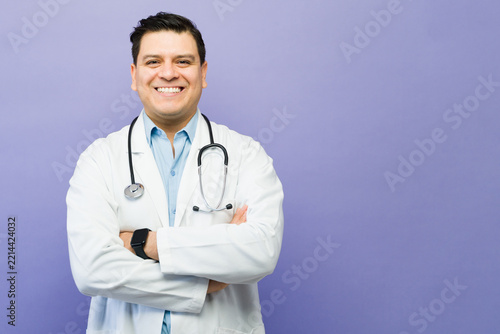 Male doctor smiling with crossed arms wearing a lab coat and stethoscope against a purple background