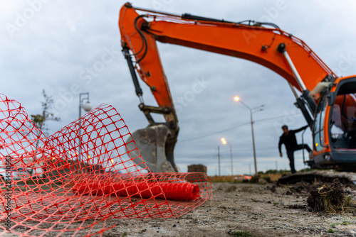 An orange hydraulic excavator lowers its bucket beside a worker at a muddy construction site, with red safety mesh in the foreground.