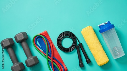 Flat lay composition of fitness equipment including dumbbells, resistance bands, jump rope, towel, and shaker bottle on a bright turquoise background with copy space for home workout themes.