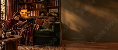 Young woman nestled on a deep green velvet sofa, engrossed in reading a book while covered by a patterned throw blanket, a small dog rests its head on the sofa beside her, cozy burrowcore style living
