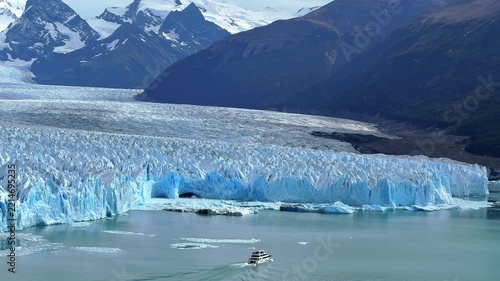 Aerial view of a boat sailing on turquoise water near the massive Perito Moreno Glacier in Patagonia, Argentina, with snow capped mountains in the background