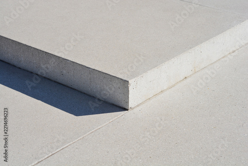 Architectural detail showing a minimalist light grey concrete step block casting a sharp shadow onto a smooth pavement surface with precise joint lines.