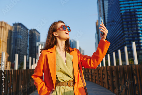 Stylish young woman in orange coat and sunglasses takes a selfie with smartphone against modern city skyscrapers. Concept of urban lifestyle, fashion and travel.