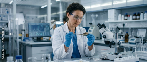 Female scientist examining sample in laboratory beaker with microscope and computer in background