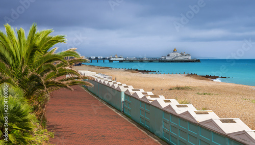 Eastbourne Pier and promenade viewed past beach huts and palm tree, East Sussex, UK