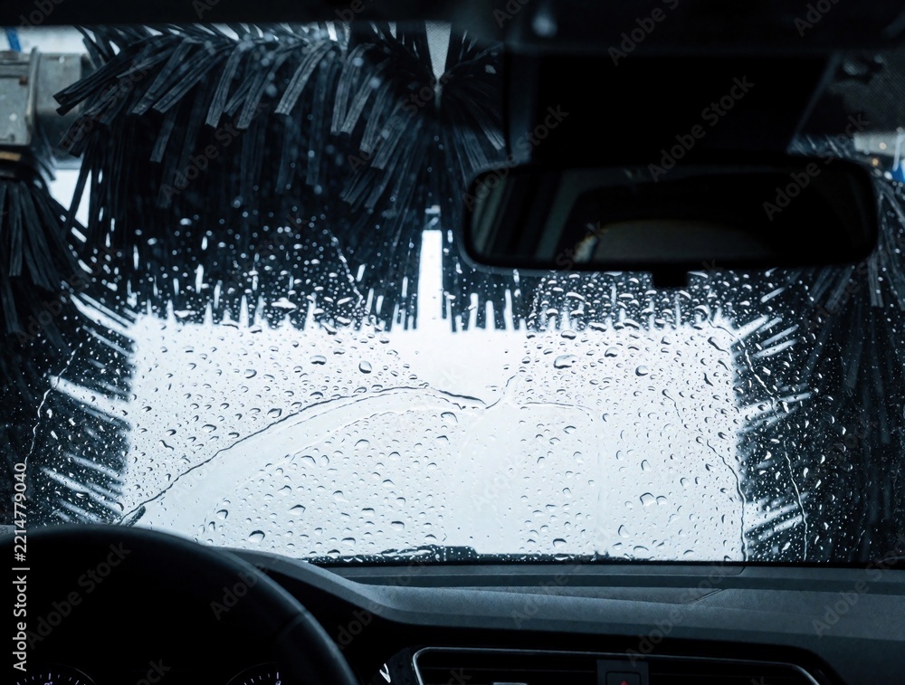 Fototapeta premium Interior view from inside car during automatic wash. Large cleaning brushes rotate outside windshield with soap suds, water droplets. Automated carwash machine cleans vehicle surface in service