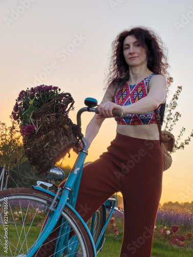 Beautiful curly-haired woman holding a blue vintage bicycle with a flower basket in a countryside field at golden hour sunset, posing in stylish crop top and trousers.