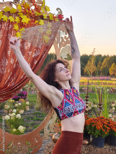 Graceful adult woman raising hands holding decorative leaf ornament while stretching outdoors in autumn field park setting with orange chrysanthemums and wooden arch backdrop.