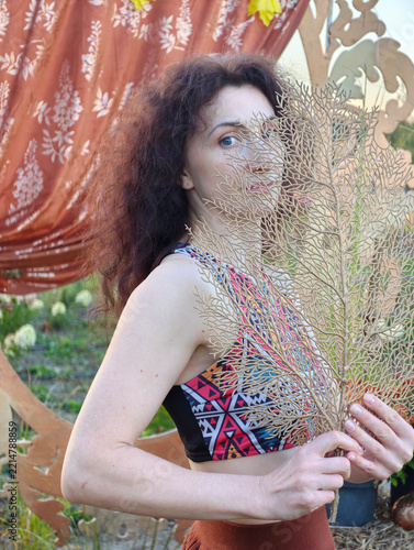Artistic portrait of a young woman holding a golden dried sea fan plant in front of her face while wearing a colorful bohemian pattern crop top outdoors in autumn setting.