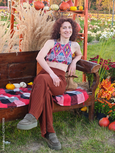 Smiling woman with curly hair relaxes on a wooden bench amidst autumn decorations, wearing a patterned top and brown wide pants, enjoying a bright fall harvest festival setting outside.