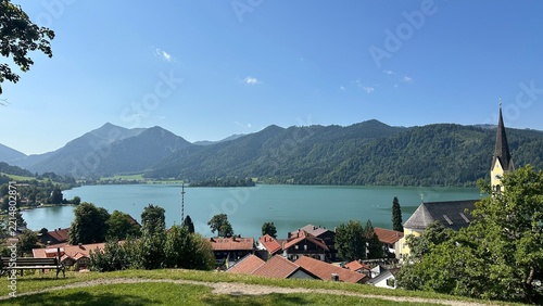 Panoramic alpine lake view with turquoise water, village rooftops, yellow church steeple, forest mountains, and under bright clear blue sky. Schliersee