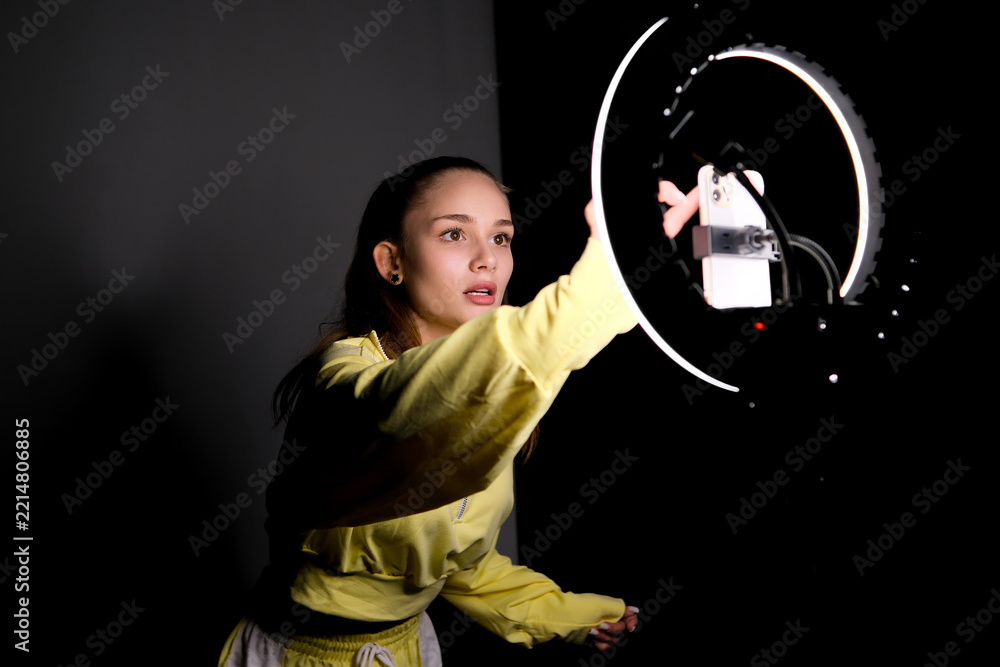 Fototapeta premium Young female content creator recording a social media video with smartphone mounted on a ring light, looking into camera in a dark home studio for livestreaming and vlogging