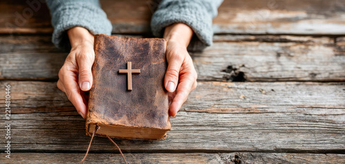 Hands gently hold a worn brown Bible with a cross on its cover above a rustic wooden table, Christian faith, spiritual devotion, prayer practice, religious study, sacred tradition, personal belief.