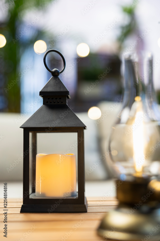 Fototapeta premium Close-up shows a black candle lantern and oil lamp glowing on a wooden rooftop table near comfortable couch seating, creating minimalist balcony ambiance.