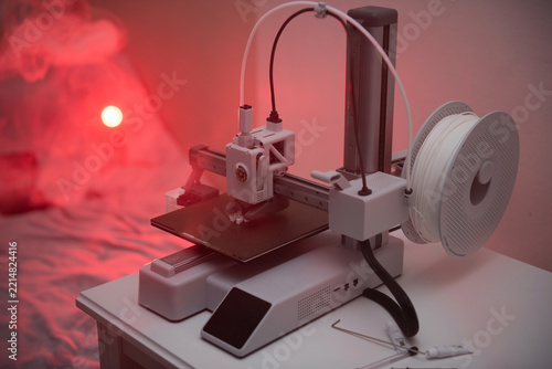 A compact white desktop 3D printer prints a detail on a table in a dark room with dramatic red backlighting, creative hobby and modern technology concept.