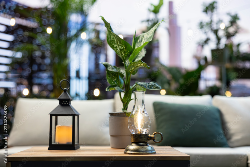 Fototapeta premium Oil lamp and lantern with a leafy potted plant sit on a wooden table beside a cushioned rooftop sofa, with blurred urban lights creating a cozy balcony setting.