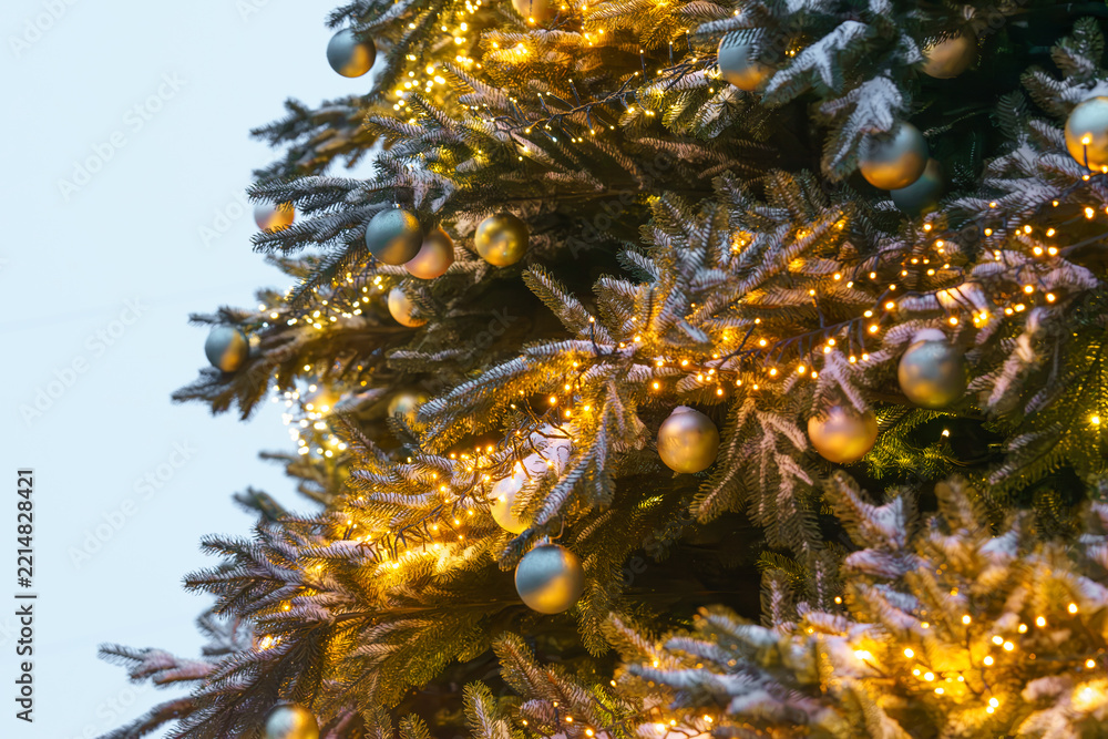 Fototapeta premium Christmas tree decorated with gold and silver ornaments and warm glowing string lights close up, creating festive holiday atmosphere against a soft blue evening background. Street, outside decorations