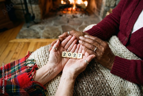 Elderly couple hands holding 2027 number tiles by fireplace on knit blanket, symbol of lasting love