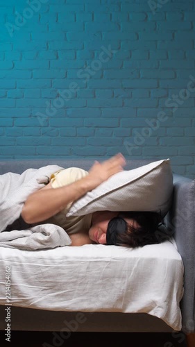 Vertical video man with a sleep mask trying to block out noise and light by covering his head with a pillow and blanket, experiencing discomfort and difficulty resting