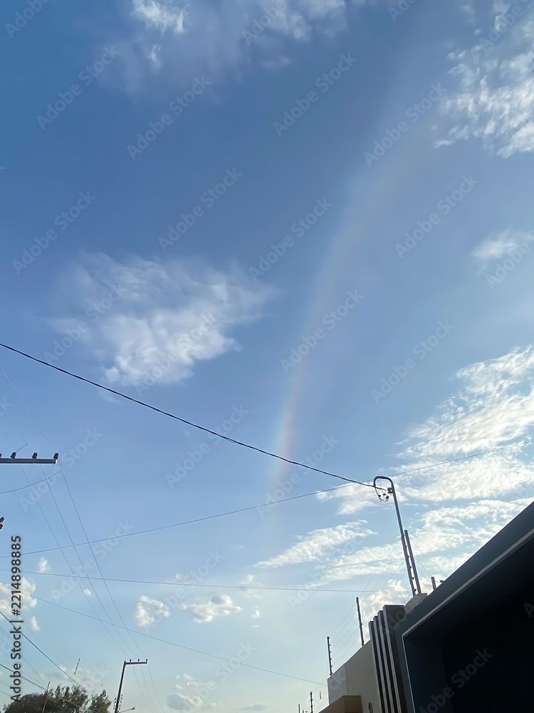 Fototapeta premium A faint rainbow arcs across a bright blue sky with scattered clouds and overhead wires