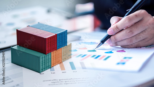 Logistics performance analysis and shipping cost management concept. Colorful miniature cargo containers with businessman writing on freight charts and manufacturing reports.