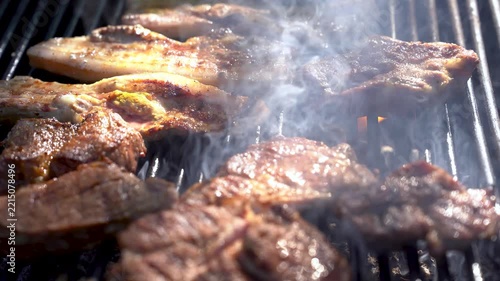 Juicy pork steaks sizzling on hot barbecue grill grates outdoors with thick aromatic smoke rising in bright warm sunlight during a summer backyard picnic cookout. 4K video.
